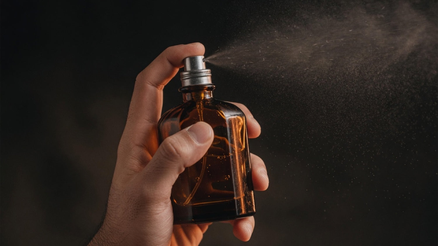 Hand spraying an unlabeled amber cologne bottle, fine mist hanging in warm light against a dark background