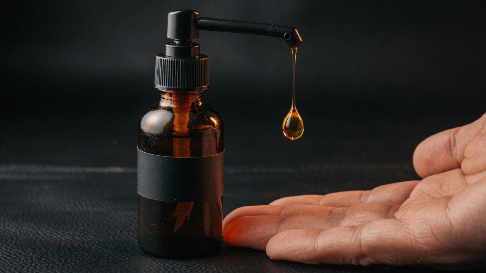 Glass dropper releasing a drop of grooming oil onto the back of a man's hand — beard oil as a multi-use men's skin moisturizer