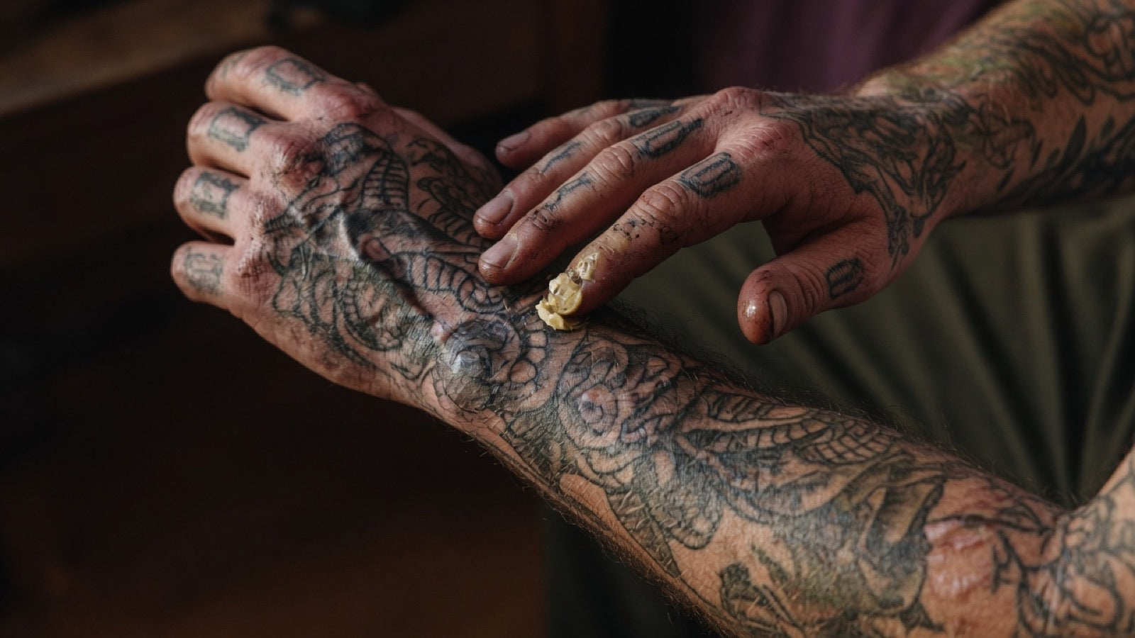Close-up of heavily tattooed hands as a fingertip applies a dab of balm to inked forearm skin in low warm workshop light