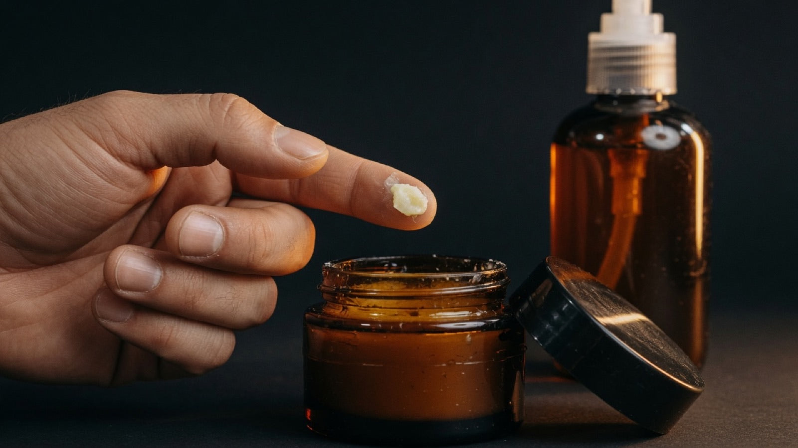 Open amber glass jar of beard balm with black lid beside it, a fingertip lifting a small amount, warm studio light