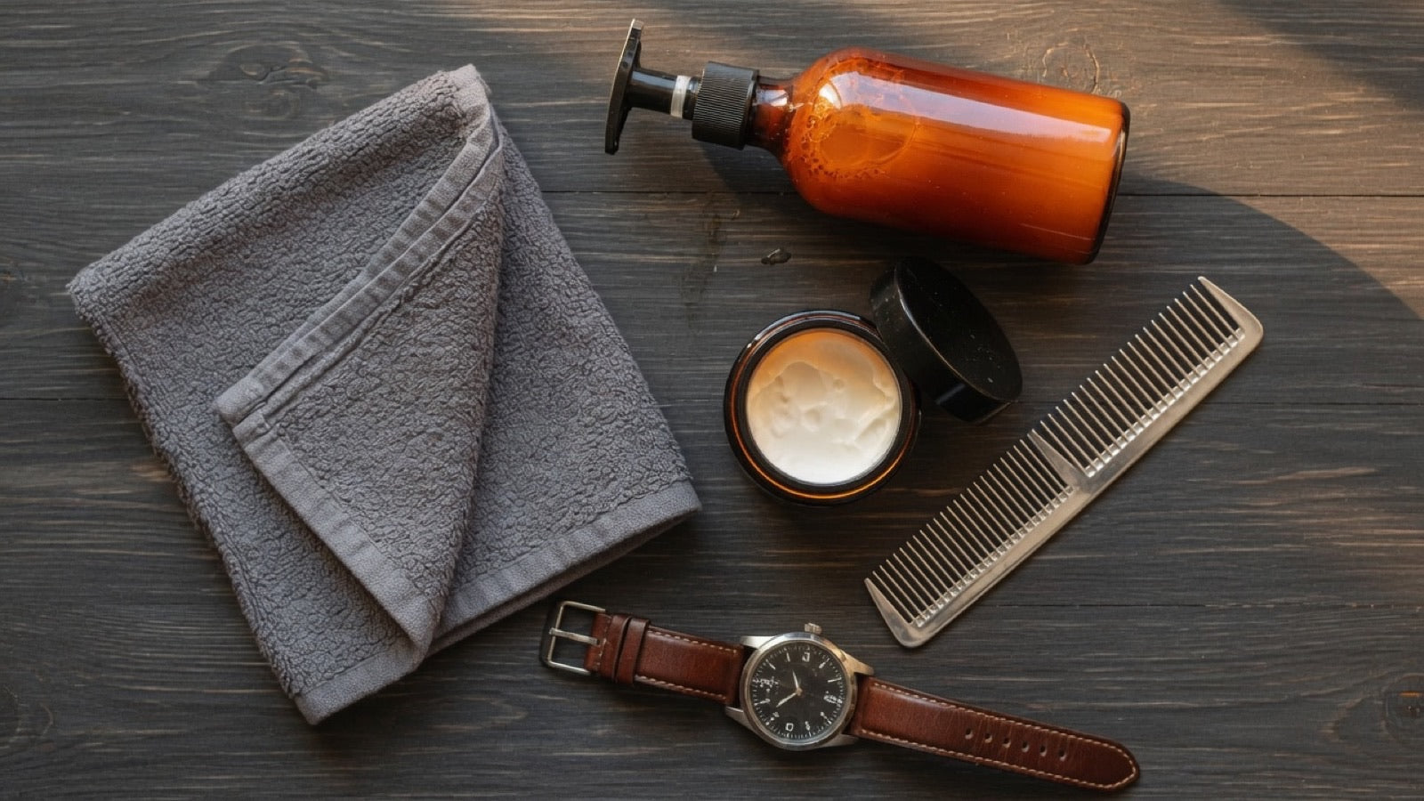 Men's skin care routine on a dark bathroom counter at dawn — towel, amber bottles, morning light