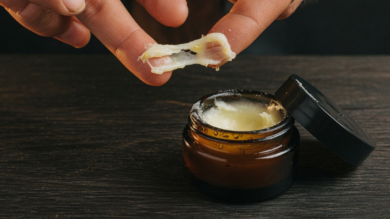 Thumb swiping a curl of beard balm from an open tin, waxy texture lifting — what beard balm does